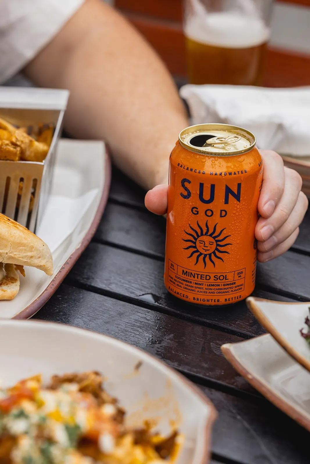 A hand holding a can of SUNGOD Minted Sol above a table of food at sunset