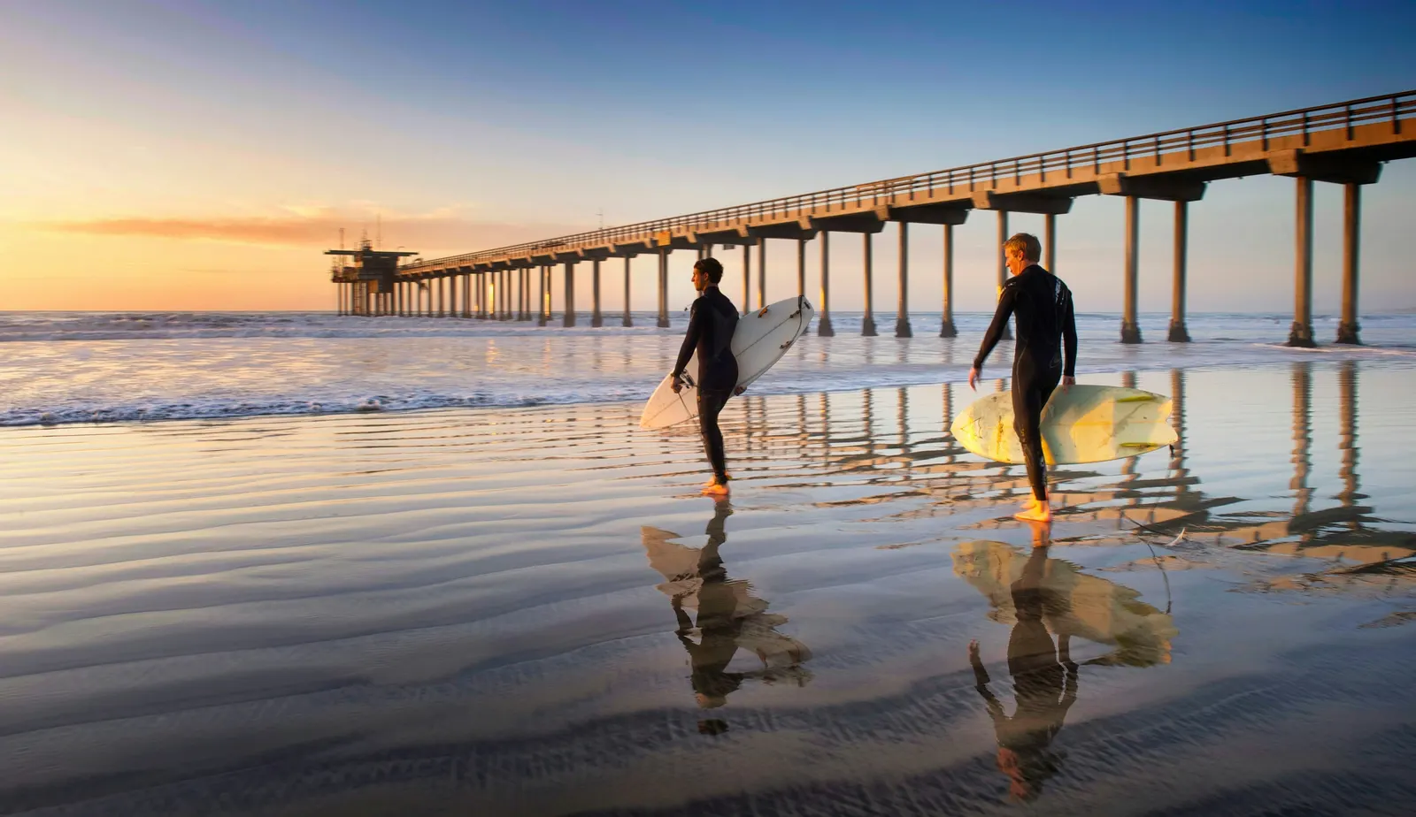 Surfers crossing the wet sand near Scripps Pier at golden hour